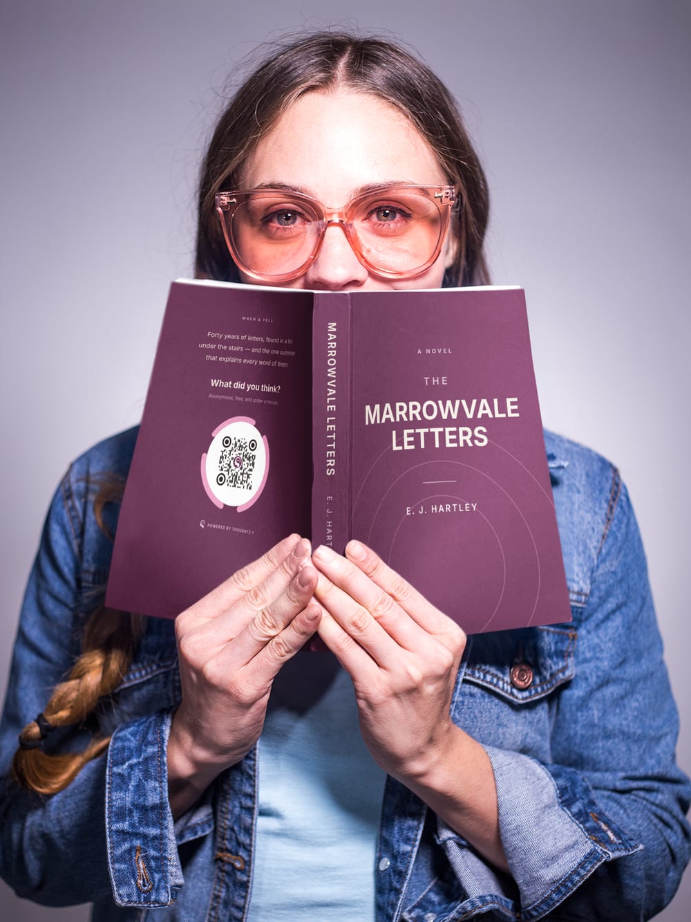 A person holding an open book up in front of their face, a Thoughtz Code printed on the back cover.